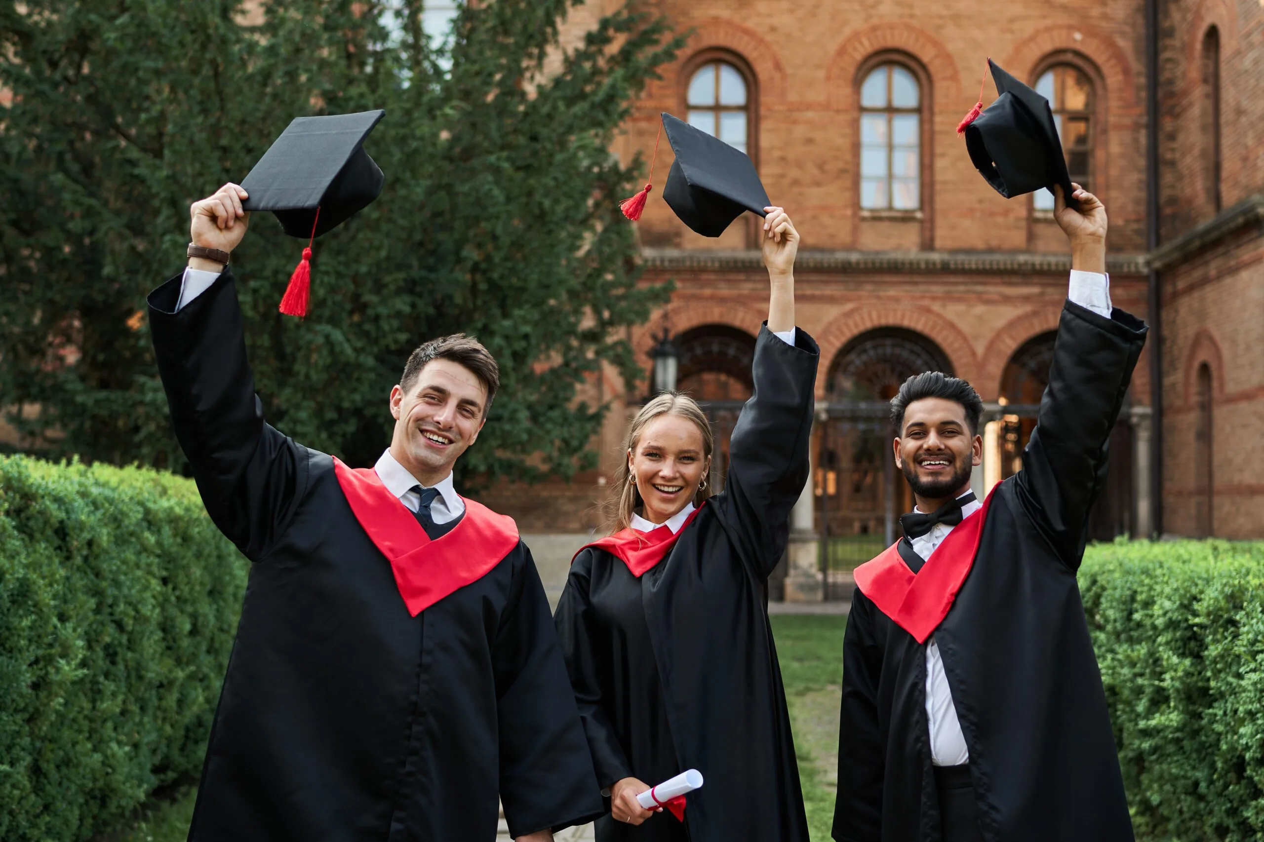 Three graduates in gowns raising their caps in front of a campus building