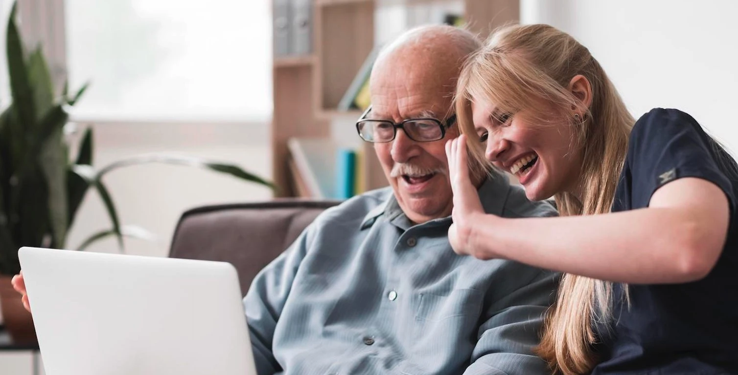 A caregiver laughing with an older man as they wave at a laptop video call
