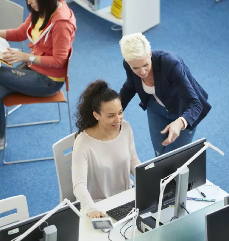 An instructor leaning in to help a student working at a computer in a library